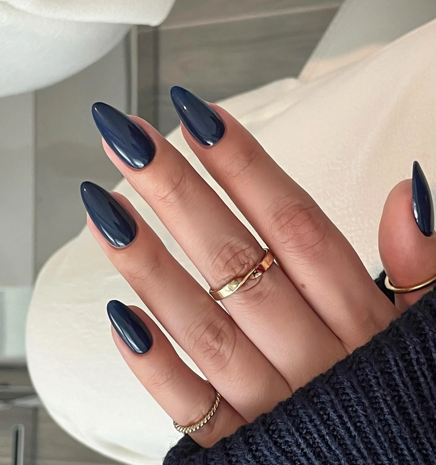 39 Navy Blue Nail Ideas That Are Too Gorgeous Not to Try in 2026 10 Glossy navy almond nails shown with gold rings on the hand. Soft natural lighting, elegant simple background.