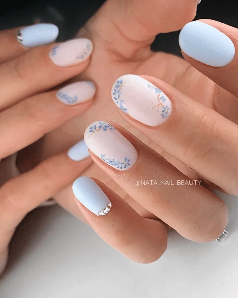 Blue and White Nail Designs 2025: 20 + Chic Ombre, French & Seasonal Ideas 13 Simple white nails with pastel blue accents and delicate floral nail art, featuring a matte finish and crystal embellishments for an elegant spring manicure.