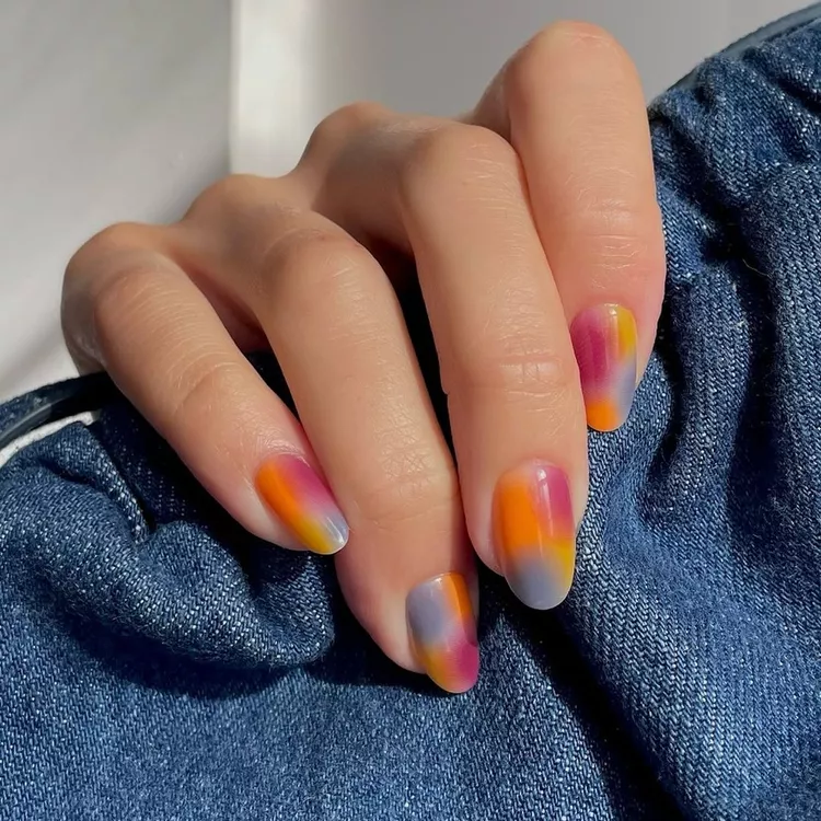 51 Easy Nail Designs You Can Do at Home (Even if You’re a Total Beginner) 49 Hand resting on denim, displaying short, oval nails with a soft, blended abstract or 'smush' design in warm autumnal colors: pink, orange, yellow, and grey.