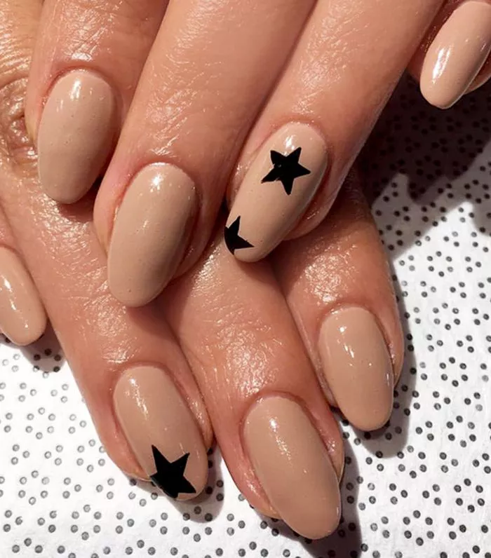 51 Easy Nail Designs You Can Do at Home (Even if You’re a Total Beginner) 47 Hands with oval nails painted a solid nude beige color, featuring one or two simple black star decals on the ring and middle fingers.