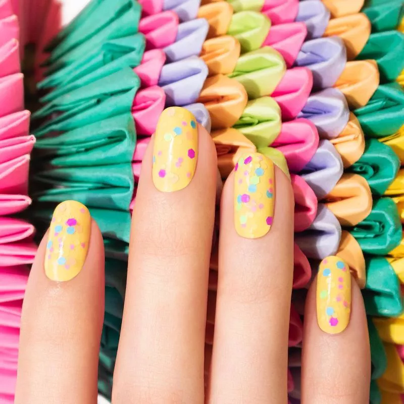 51 Easy Nail Designs You Can Do at Home (Even if You’re a Total Beginner) 32 Fingers with short, oval nails painted a solid bright yellow and covered in a topcoat containing multicolored confetti glitter dots (pink, blue, mint green) against a colorful woven background.