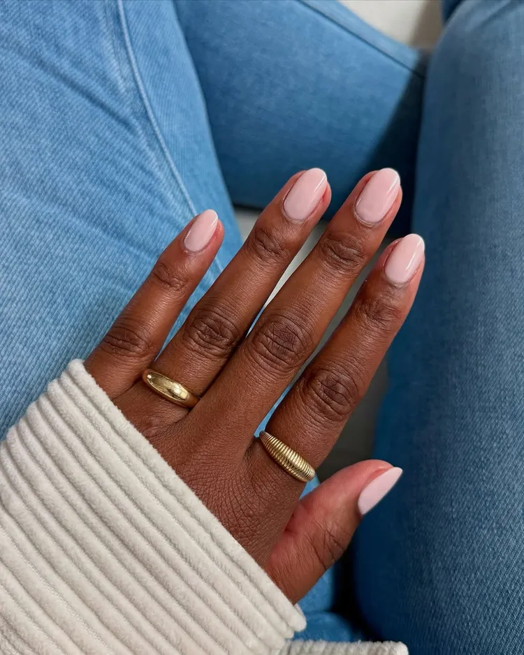 32+ Best Nail Colors for Dark Skin That Look Absolutely Stunning 14 A hand with deep brown skin wearing a subtle, dreamy pastel pink nail polish with a glossy finish. The hand is relaxed against blue jeans and wearing a cream knit sweater and gold rings.
