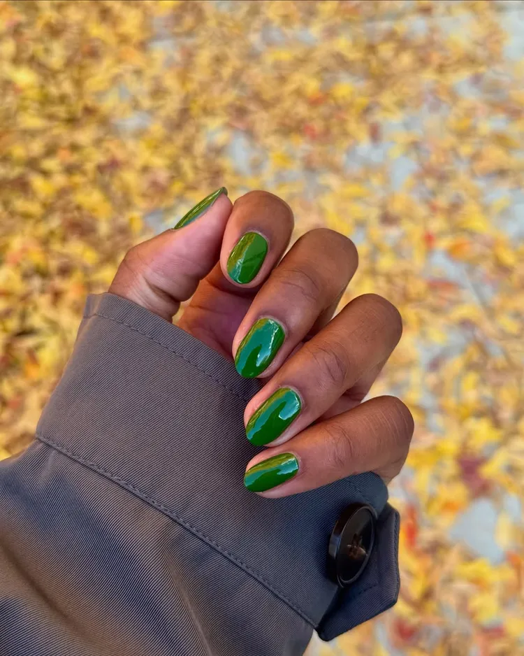 32+ Best Nail Colors for Dark Skin That Look Absolutely Stunning 38 Hand with deep brown skin and a gray jacket sleeve, featuring short, glossy oval nails painted in a rich, opaque enchanting olive green polish. The background is a blurred field of yellow autumn leaves.