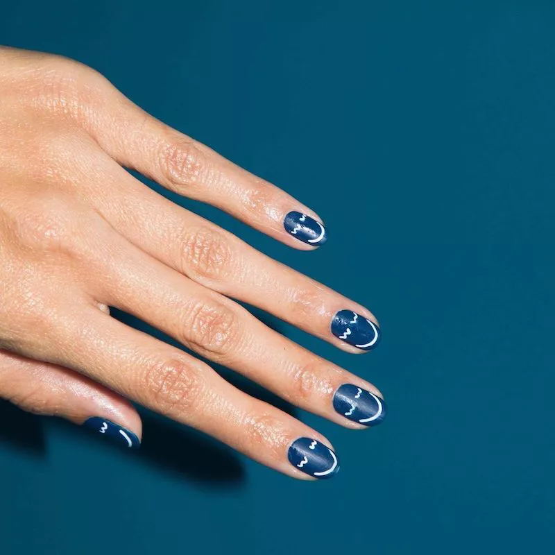 51 Easy Nail Designs You Can Do at Home (Even if You’re a Total Beginner) 37 Close-up of short, rounded nails with a solid deep navy blue base, featuring a simple, white line art design resembling a stylized frown or smile face on each nail.