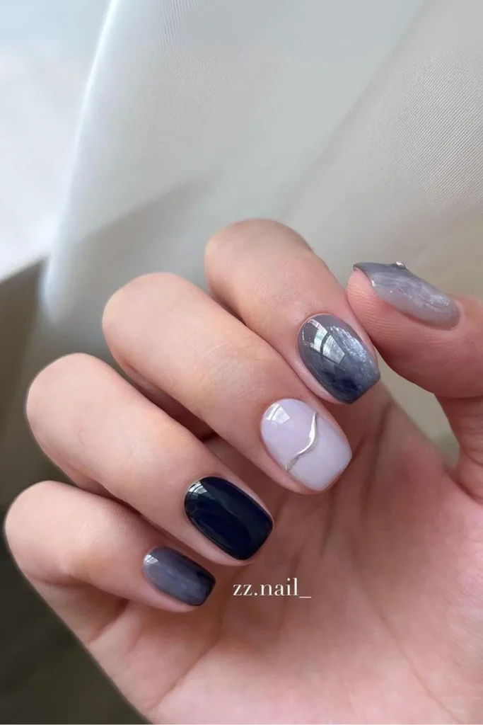 Short square dark grey and navy marble nails mixed with pale lavender polish and a thin silver swirl line