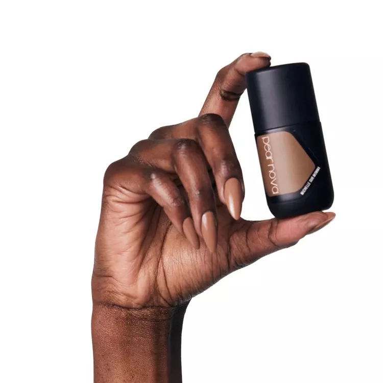 32+ Best Nail Colors for Dark Skin That Look Absolutely Stunning 35 Hand with deep brown skin holding a matte black bottle of Dearnova nail polish. The short, almond-shaped nails are painted in a creamy, glossy milk-chocolate brown nude polish, perfect for all skin tones