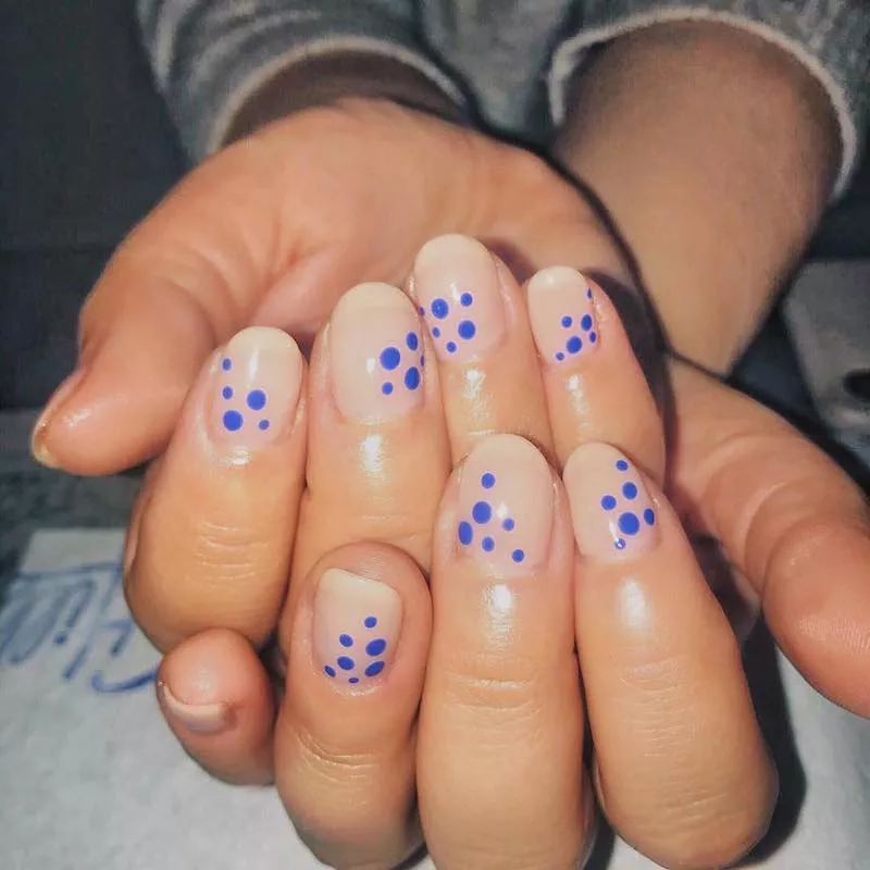 51 Easy Nail Designs You Can Do at Home (Even if You’re a Total Beginner) 17 Close-up of hands with round, milky nude nails featuring a delicate, randomized pattern of small cobalt blue polka dots over the center of the nail bed.