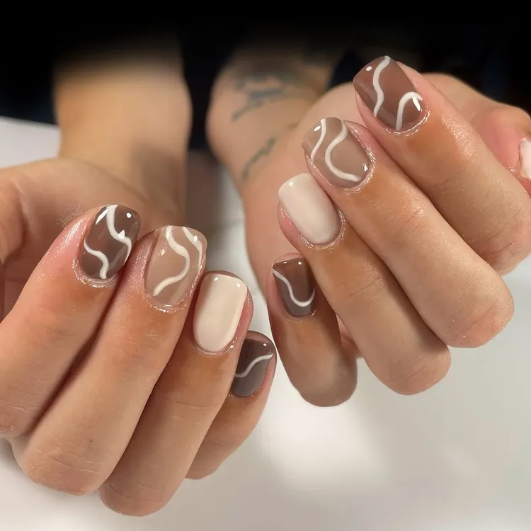 51 Easy Nail Designs You Can Do at Home (Even if You’re a Total Beginner) 50 Hands with short, oval nails featuring a mix of solid earth-toned nude and brown polishes, with some nails accented by a white abstract swirl design.