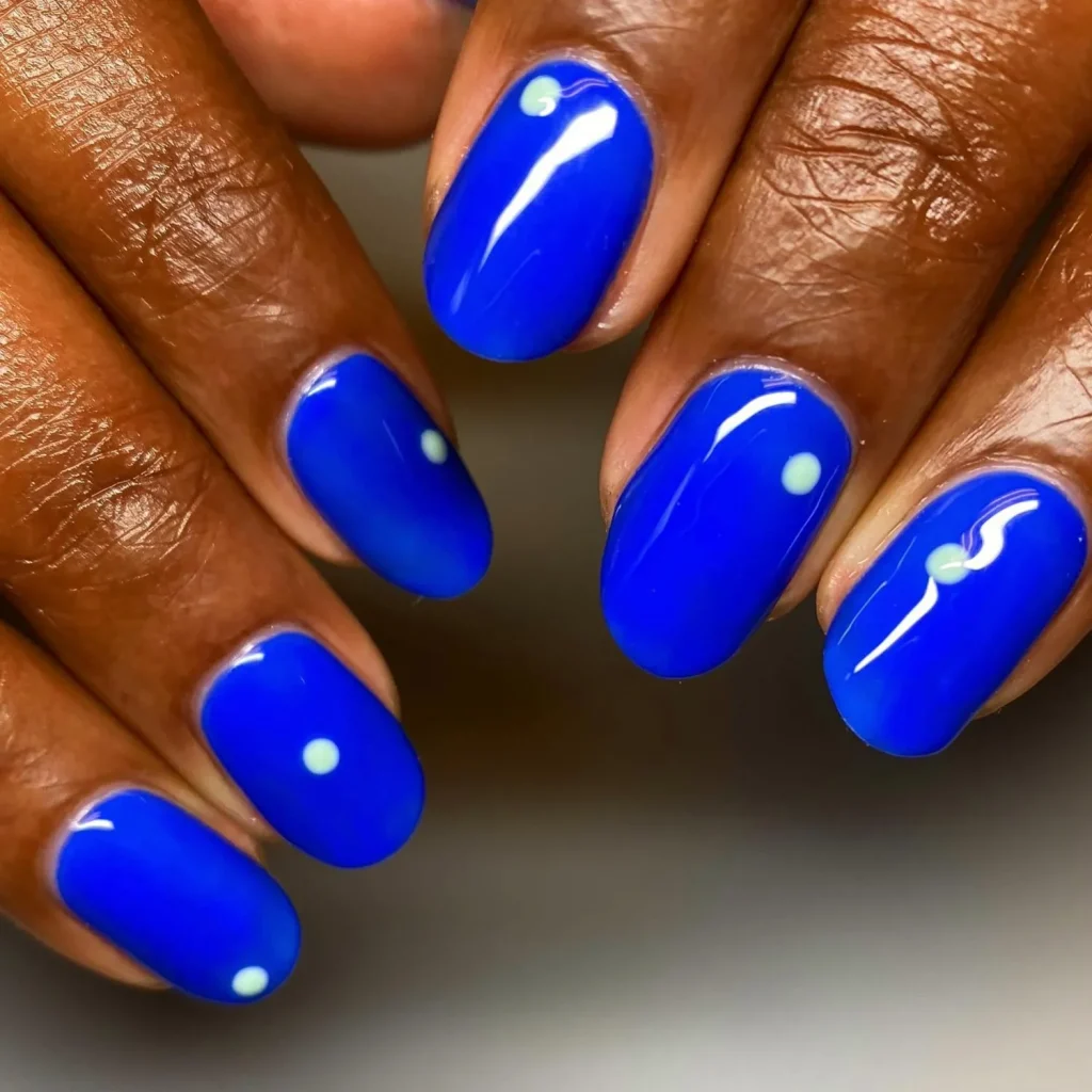 51 Easy Nail Designs You Can Do at Home (Even if You’re a Total Beginner) 15 Close-up of hands showing vibrant, glossy electric blue oval nails with a single small, off-center white dot on each nail.