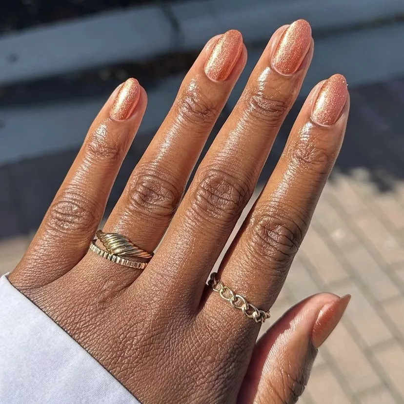 32+ Best Nail Colors for Dark Skin That Look Absolutely Stunning 8 Close-up of short, oval-shaped nails on deep brown skin painted in a shimmering metallic rose-gold or copper-orange polish. The hand is wearing gold rings and a white shirt cuff, glistening in the sunlight.