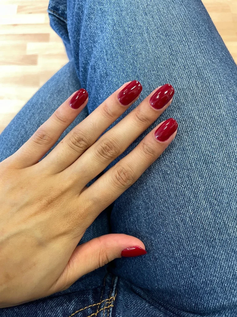 Nail Shapes for Wide Nail Beds: Flattering Styles to Slim Your Fingers 3 A close-up view of a hand resting on denim jeans, showing oval-shaped nails painted with a deep, glossy red polish.
