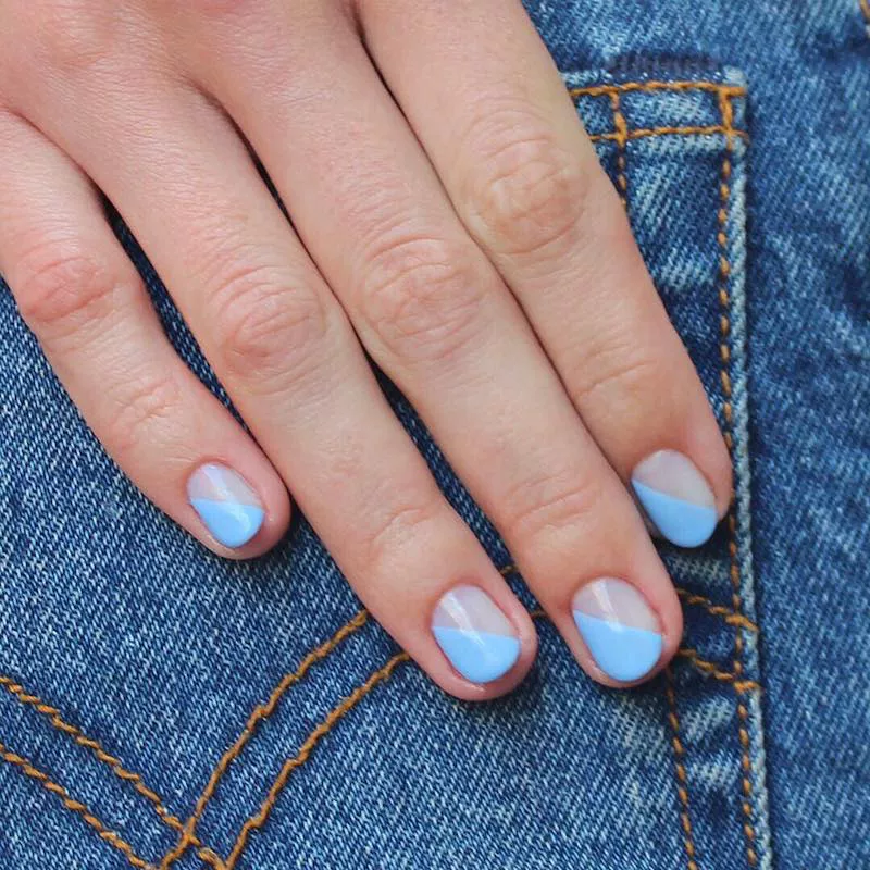 51 Easy Nail Designs You Can Do at Home (Even if You’re a Total Beginner) 20 Hand resting on a denim lap, displaying short, clear nails with a negative space design where a block of baby blue or periwinkle polish covers the tip in a clean diagonal line.