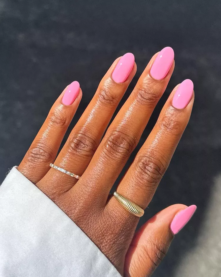 32+ Best Nail Colors for Dark Skin That Look Absolutely Stunning 10 Close-up of almond-shaped nails painted in a bright, glossy bubblegum pink polish on deep brown skin. A hand wearing a white sleeve and thin gold rings is featured.