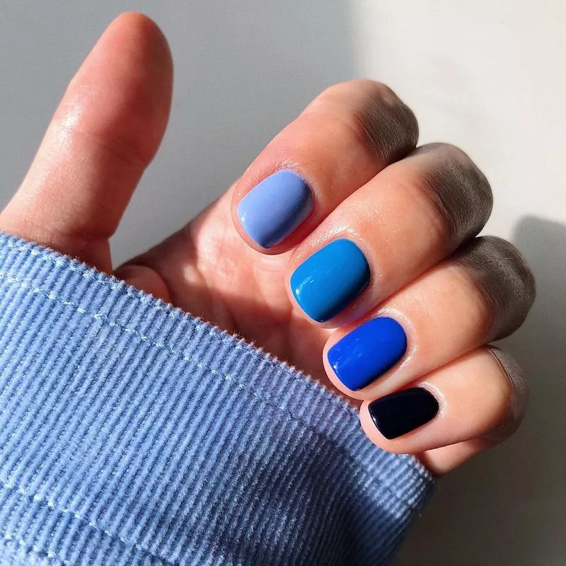 51 Easy Nail Designs You Can Do at Home (Even if You’re a Total Beginner) 35 Hand emerging from a light blue shirt cuff, showing a gradient manicure where each short, square nail is painted a different shade of blue, transitioning from pale periwinkle to deep navy.