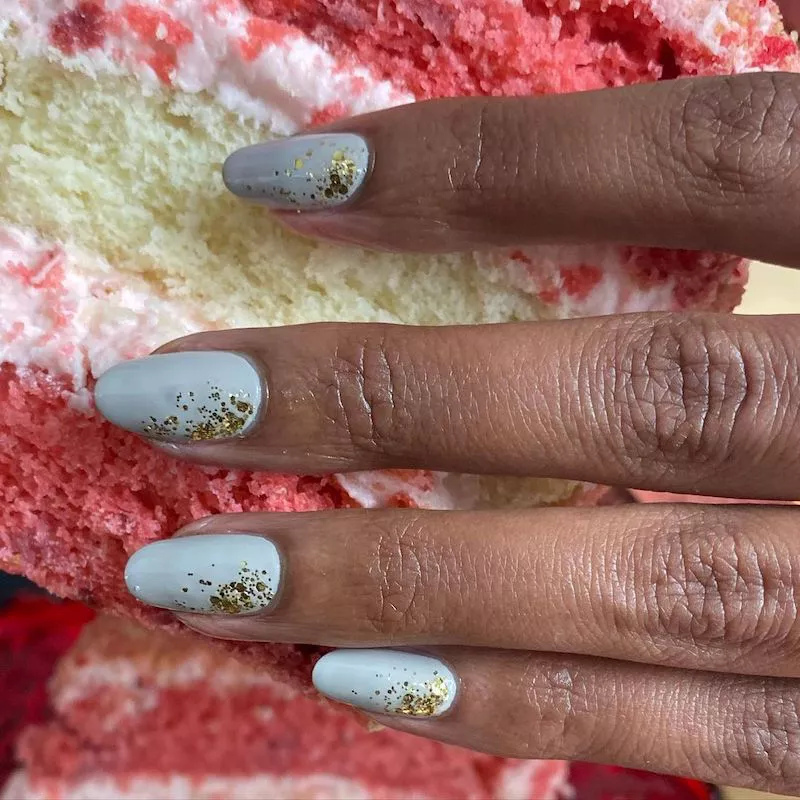 51 Easy Nail Designs You Can Do at Home (Even if You’re a Total Beginner) 19 Close-up of fingers holding a slice of cake, with light gray or dusty blue oval nails featuring chunky gold glitter concentrated around the cuticle area, fading out toward the tip.