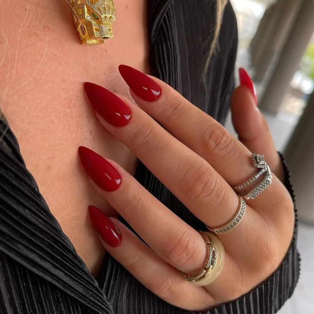 Nail Shapes for Wide Nail Beds: Flattering Styles to Slim Your Fingers 6 A hand wearing three ornate gold and diamond rings, showing long, glossy, bright red stiletto-shaped nails.
