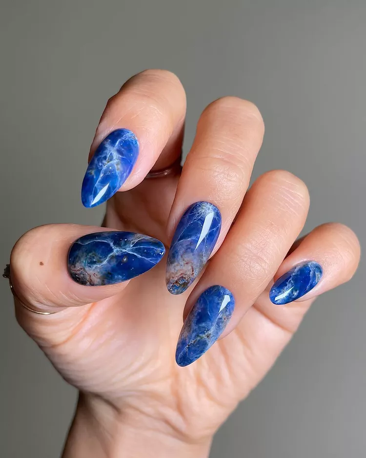 30 Stunning Dark Blue Nail Designs for a Denim-Inspired Summer Look 2 Glossy Lapis Lazuli gemstone nail art on long almond-shaped nails. Features a deep royal blue marble effect with white vein details and earthy brown and gold accents. A luxurious and trendy stone-effect manicure idea.